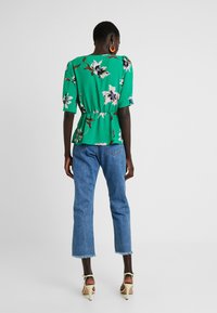 Blusa peplum verde con flores y mangas cortas; combinada con jeans cortos de color azul claro y sandalias de tacón color beige. Patrón floral de alto contraste.