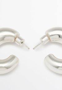 Boucles d'oreilles en argent en forme de créoles, présentant un design épais et courbé avec une finition polie. Les tiges sont visibles au centre pour assurer le maintien dans l'oreille.