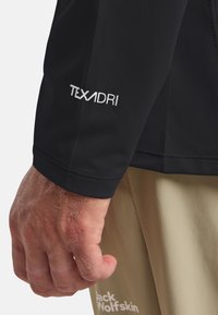 Schwarzes Langarmshirt mit TEXADRI-Logo am Ärmel; glatte Textur mit enganliegenden Manschetten. Hand sichtbar, trägt hellkhakifarbene Hosen.