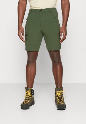 Mann trägt olivgrüne Cargo-Shorts mit Reißverschluss und braune Wanderschuhe mit gelben Schnürsenkeln, steht vor einem schlichten hellen Hintergrund.
