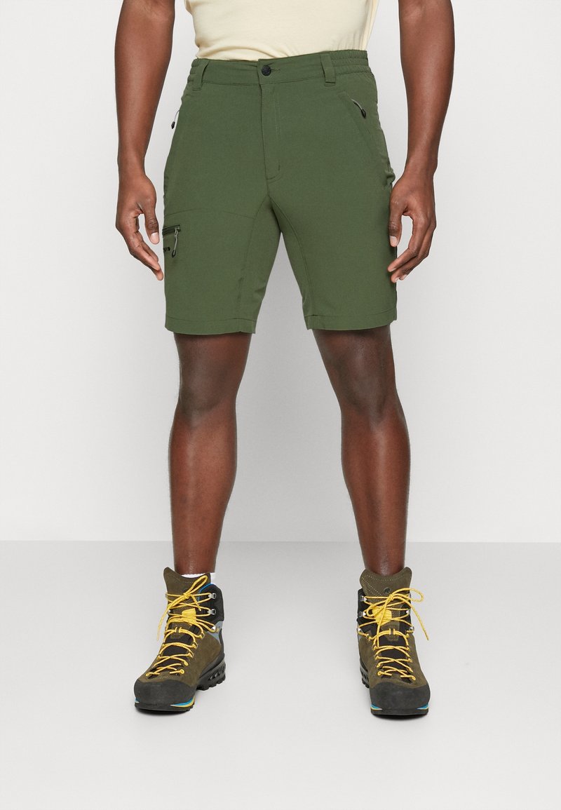 Homme portant un short cargo vert olive à fermeture éclair et des chaussures de randonnée marron avec des lacets jaunes, debout sur un fond clair uni.