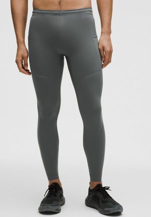 Hombre vistiendo leggings deportivos grises ajustados y zapatillas negras para correr, de pie contra un fondo claro y sencillo.