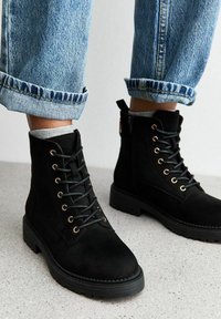 Svart mocka ank stövlar med rund tå, prydda med guldöglor och svarta snören. Sulan är tjock och texturerad; parat med uppvikta jeans.