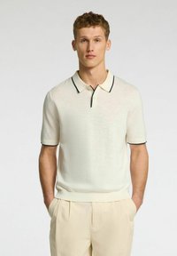 Polo shirt corto crema a maniche corte con una trama fine, caratterizzato da un collo scuro a contrasto e dettagli sulle maniche, abbinato a pantaloni leggeri beige.