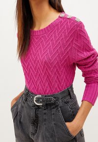 Heldere roze gebreide trui met een visgraatpatroon, voorzien van decoratieve bloemenknopen op de schouder en lange geribbelde mouwen. Gecombineerd met hoge zwarte jeans die een gestikte riem laten zien.