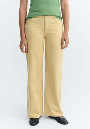 Person trägt eine hochtaillierte, weit geschnittene, beige Cordhose, ein grünes ärmelloses Oberteil und schwarze flache Schuhe und steht vor einem schlichten Hintergrund.