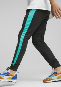 Black athletic pants with a turquoise side stripe, featuring a slim fit and elastic cuffs. Worn with multicolored sneakers.
