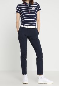 Polo rayé bleu marine et blanc avec col zippé et logo, associé à un pantalon bleu marine et des baskets blanches. La chemise est ajustée, avec des manches courtes.
