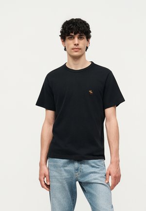 Jeune homme aux cheveux bouclés et foncés portant un T-shirt noir avec un petit logo animal brun sur la poitrine et un jean bleu clair, debout.