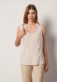 Beige ärmlös topp med rundade halsringning och mjuk textur, parat med ljusbeige byxor. Modellen håller en matchande hoodie.