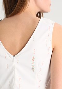 Top sans manches blanc présentant un dos en V, un motif multicolore subtil et un petit zip au niveau de l'encolure arrière. Texture de tissu lisse.