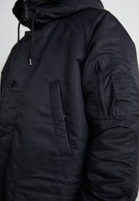 Schwarze Pufferjacke aus glattem, glänzendem Material. Mit Kapuze, vorderen Knöpfen, seitlichen Taschen und einer Reißverschlusstasche am Ärmel.