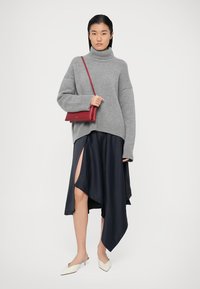 Siva pletena turtleneck pulover, asimetrična temno modra satenska krila z visokim razporkom, v kombinaciji z rdečo torbico prek telesa in belimi mule sandalami.