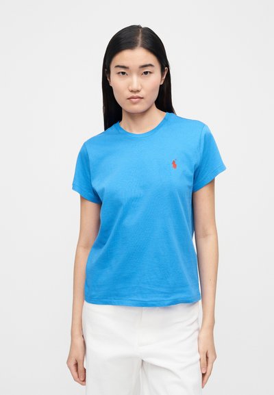 Mėlyna medvilninė marškinėliai su trumpomis rankovėmis, apvalia iškirpte ir mažu raudonu logotipu ant krūtinės. Lygus audinys ir laisvas siluetas.