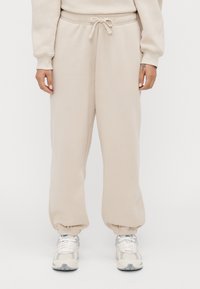 Persoon in beige sweatpants met elastische boorden en wit-grijze sneakers staat tegen een effen achtergrond.