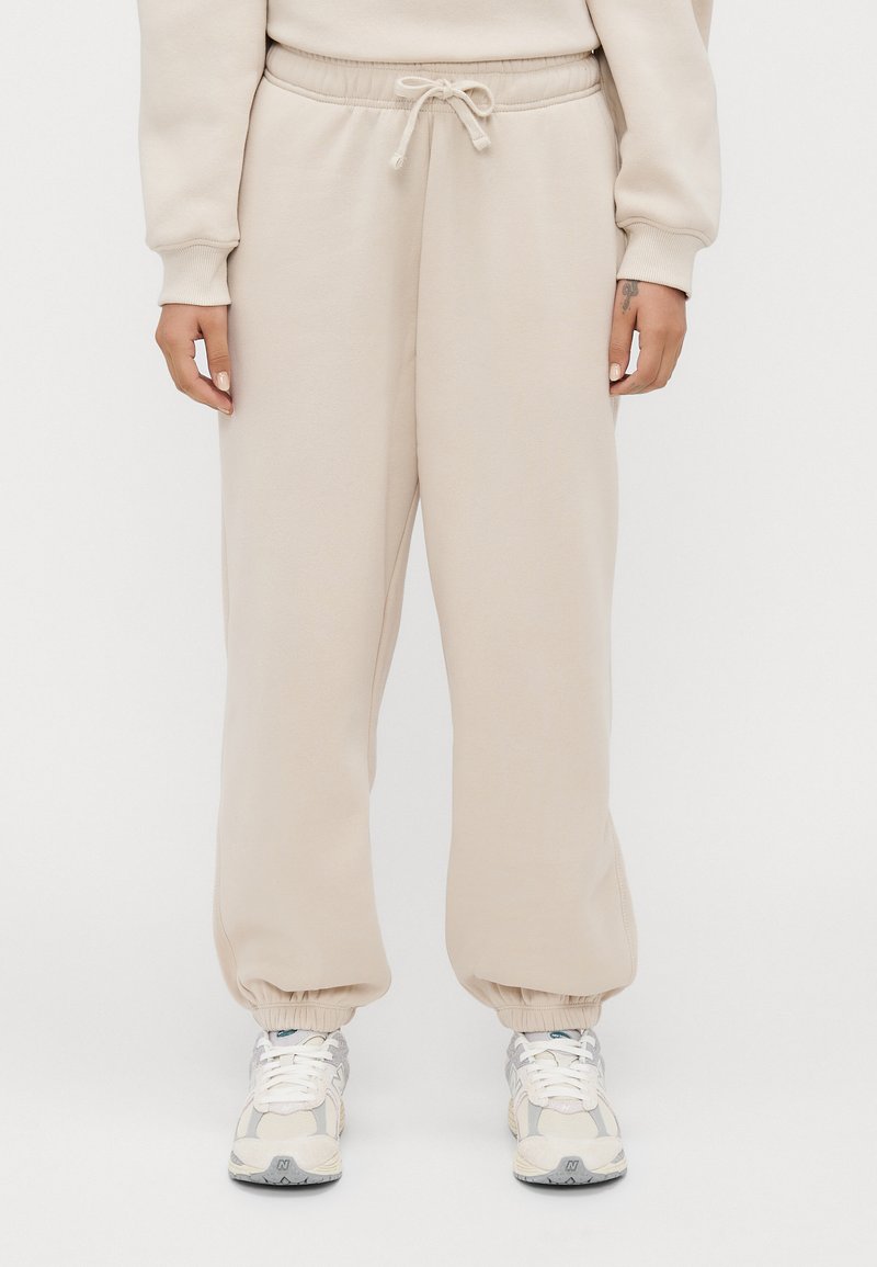 Persoon in beige sweatpants met elastische boorden en wit-grijze sneakers staat tegen een effen achtergrond.