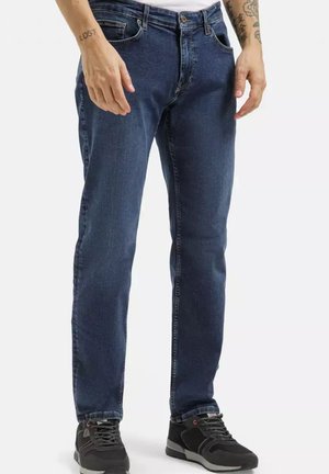 Mężczyzna noszący ciemnoniebieskie jeansy o prostym kroju i czarne trampki, stojący z rękami swobodnie opuszczonymi wzdłuż ciała na białym tle.