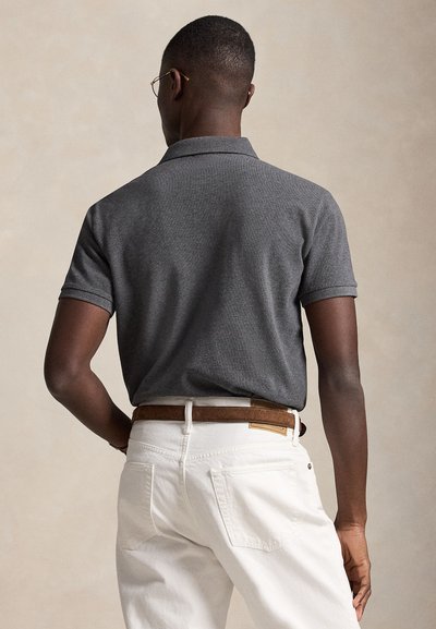 Homme portant un polo gris à manches courtes, un pantalon blanc et une ceinture marron, debout, dos à la caméra.