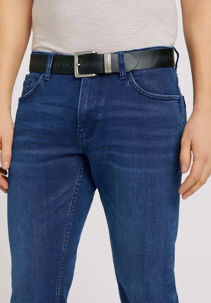 Sort læderbælte med en sølvmetalkrog, båret over mørkeblå denimjeans med en subtil tekstur og klassisk fem-lomme design.