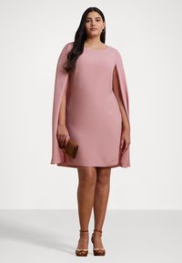 Vestido sin mangas rosa con diseño de capa, hecho de un tejido suave. Presenta un escote redondo y un dobladillo fluido. Combinado con un bolso de mano marrón y tacones.