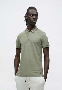 Oliwkowa koszulka polo z usztywnianym kołnierzykiem, patką na trzy guziki i subtelnym haftem logo. Noszona z jasnoszarymi dresami.