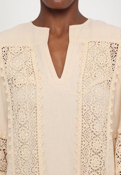 Beige tunic made of lightweight fabric featuring intricate lace detailing on sleeves and sides, with a V-neck and decorative trim.