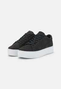 Ein Paar schwarze Schnürsneaker mit dicken weißen Sohlen auf einem schlichten hellgrauen Hintergrund, leicht nach rechts geneigt.