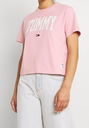 Kobieta ubrana w jasnoróżowy t-shirt Tommy Jeans z białym haftowanym logo i jasnoniebieskie, wysokie dżinsy, stojąca na jednolitym tle.