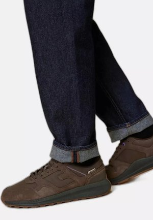 Persona con vaqueros azul oscuro con puños y zapatillas marrones, dando un paso adelante con un pie sobre un fondo blanco.