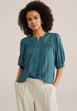 Žena s kučeravými vlasmi, oblečená v tyrkysovej blúzke s čipkou a béžových vysokých nohaviciach, s rukami v vreckách, neutrálne pozadie.