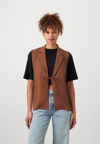 Bruin linnen vest met een geknoopte sluiting aan de voorkant, een reverskraag en open zijkanten, gecombineerd met een zwart cropped t-shirt en lichtblauwe denim jeans.