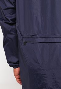 Gros plan sur le bras et la main d'une personne portant une veste légère bleu marine avec une poche latérale zippée et un ourlet élastique.