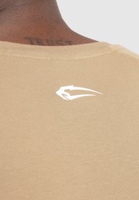 Beige bomullströja med en vit logotyp tryckt på övre ryggen. Tyget har en slät, lätt textur och rundad halsringning.