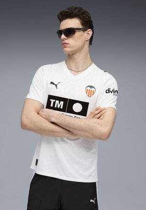 Hvid sportsjersey med et struktureret mønster, prydet med Valencia CF-logo, sort grafisk print og Puma-logo. Bæres med sorte shorts.
