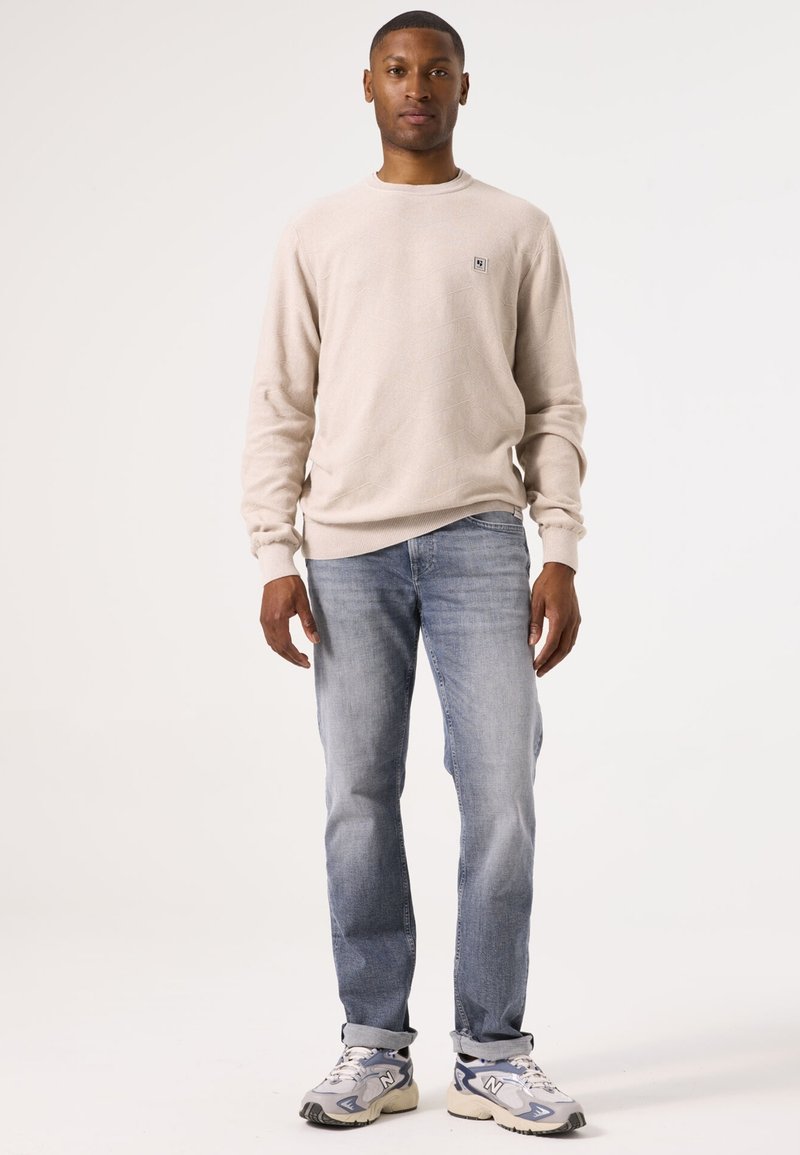 Beiger Pullover mit einem strukturierten Rautenmuster, kombiniert mit hellblauen Jeans und grauen Sneakers mit weißen Akzenten. Das Modell steht aufrecht.