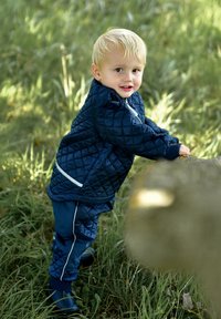 Chaqueta acolchada azul marino con detalles blancos y pantalones a juego, presentada entre hierba verde, con un niño interactuando con un árbol.