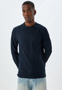 Marinblus med lång ärm och rundad halsringning, ribbad textur och avslappnad passform, ihop med ljusblå jeans. Tydlig design utan logotyper.