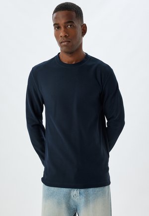 Marineblaue Langarmshirts mit Rundhalsausschnitt, gerippter Textur und lockerer Passform, kombiniert mit hellblauen Jeans. Klare Details ohne Logos.