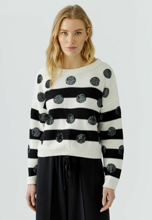 Blonde vrouw die een witte trui draagt met zwarte horizontale strepen en grote zwarte stippen, gecombineerd met een zwarte broek.