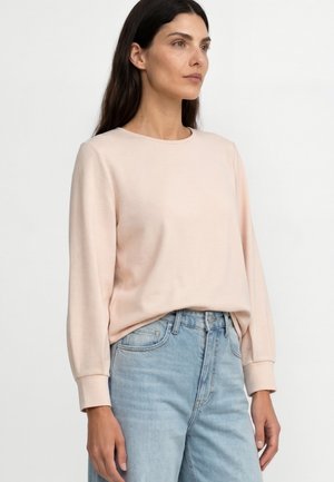 Frau trägt ein hellrosa langärmeliges Oberteil und hellblaue hochtaillierte Jeans und steht vor einem schlichten weißen Hintergrund.