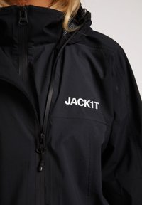 Sort vandtæt jakke med høj krave, lynlås foran og stort logo "JACK1T" på venstre bryst. Har struktureret stof og elastiske detaljer.