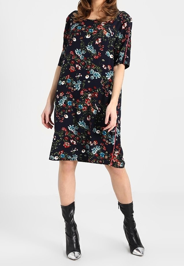 Femme portant une robe noire fleurie longueur genoux avec des manches courtes et des bottes noires mi-mollet avec embouts métalliques.