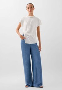 T-shirt bianco a maniche corte con dettaglio a nodo, abbinato a jeans a gamba larga di un blu chiaro. Gli accessori includono una collana voluminoso e sandali semplici.
