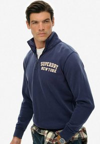 Granatowy polarowy pulower z suwakiem, z haftem "SUPERDRY NEW YORK" w jasno beżowym kolorze. Teksturowana tkanina, luźny krój.