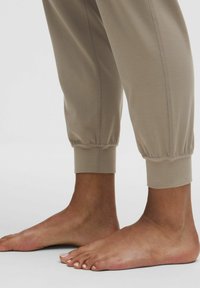Barfuß mit beigen Jogginghosen, die an den Knöcheln hochgerollt sind, auf einem hellen Hintergrund.
