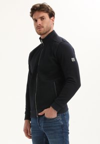 Hombre con cabello castaño claro, vestido con una chaqueta negra con cremallera y jeans azules, de pie contra un fondo blanco liso.