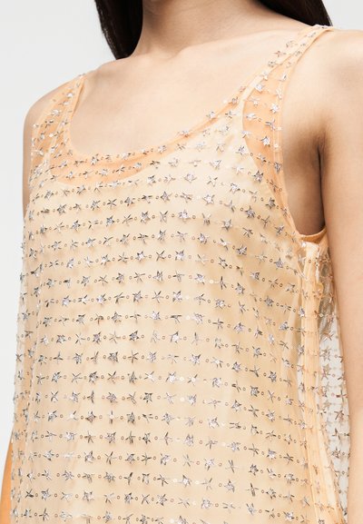 Top bege transparente sem mangas com lantejoulas prateadas em forma de estrela, usado por uma pessoa com cabelo longo e escuro, close-up no torso superior.