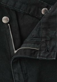Vaqueros de mezclilla negros con una cremallera visible, herrajes plateados y detalles desgastados. La textura de la tela es suave con un ligero brillo.