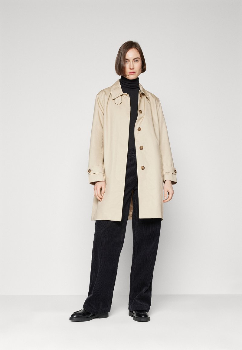 Gabardina beige con cierre de botón y cuello puntiagudo, llevada sobre un suéter de cuello alto negro. El modelo la combina con pantalones de pana negros y zapatos.