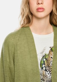 Olivgrüner Strickcardigan mit weicher Textur, der einen weit offenen Vorderteil und gerippte Details aufweist, über einem weißen Grafik-T-Shirt getragen.